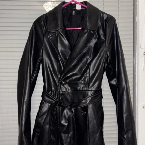 Svart trenchcoat från H&M - Snygg svart trenchcoat från H&M i syntetmaterial. Jackan har en klassisk design med knappar och ett bälte i midjan för en stilren look. Perfekt för höst och vår med sina långa ärmar och tidlösa stil. Använt endast 2 gånger. Köpt för 700kr