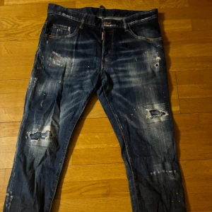 Blå jeans från Dsquared2 - Snygga äkta blå jeans från Dsquared2 med slitna detaljer och färgstänk. De har en klassisk femficksdesign och en läderpatch med logotyp på baksidan. Perfekta för en avslappnad stil. Har använts cirka 10 gånger men har legat i garderoben länge och därför är skrynklig. Är storlek 32 men har ett mindre passform så passar bäst på storlek 30.