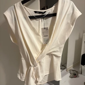 Vit omlottopp från Zara - Säljer en stilren vit omlottopp från Zara. Toppen har en v-ringad design och är ärmlös, vilket ger en elegant look. Perfekt för att klä upp eller ner beroende på tillfälle. Helt oanvänd med prislapp 