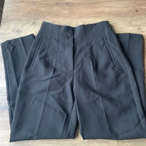 Svarta kostymbyxor - Snygga svarta kostymbyxor med hög midja och stilrena fickor. Byxorna har en diskret dragkedja och knapp framtill för en elegant look. Perfekta för en chic och professionell stil.