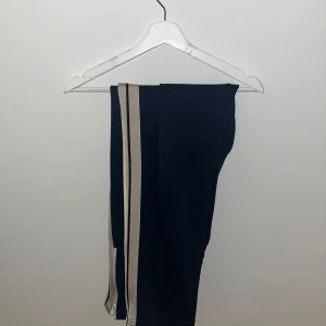 Svarta byxor med beiga ränder från Palm Angels - Snygga blåa byxor från Palm Angels med beiga ränder längs sidorna. Perfekta för en stilren look. Byxorna har en elastisk midja för extra komfort. Tveka inte vid frågor💯🔥