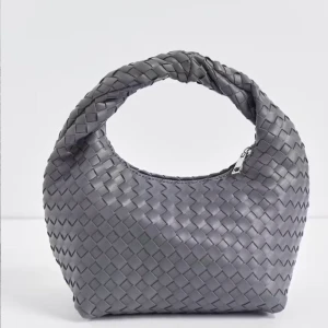 Grå flätad handväska - Snygg grå handväska med flätat mönster och dragkedja. Perfekt för att ge en stilren touch till din outfit. Väskan har en unik design med ett vridet handtag som ger extra karaktär.