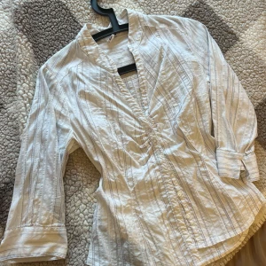 Vit randig skjorta från H&M - Snygg skjorta med ränder. Skjortan har en v-ringning och tre knappar längst ner. Ärmarna är tre fjärdedels längd. Skjortan är i ett fint, lätt material. Den är i mycket gott skick, storlek S.
