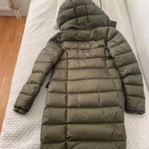 Grön dunjacka från Marc O’Polo  - Säljer en lång grön dunjacka från Marc O’ Polo. Jackan har en huva och stängs med dragkedja. Perfekt för kalla dagar med sin vadderade design.