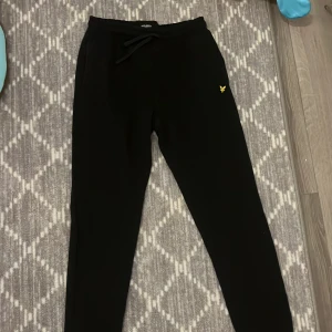 Svarta mjukisbyxor från Lyle & Scott Junior - Säljer ett par svarta mjukisbyxor från Lyle & Scott Junior. Byxorna har en elastisk midja med snörning och en liten gul logga på framsidan. De har en bakficka och ribbade muddar vid bensluten för en bekväm passform.