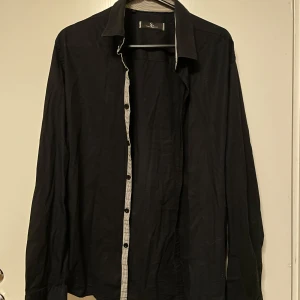 Svart skjorta från Savvy Citizen - Snygg svart skjorta från Savvy Citizen med långa ärmar och stilrena knappar. Perfekt för en elegant look. Den har en diskret randig detalj på insidan av kragen som ger en extra touch.