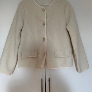 Beige teddyjacka med knappar - Mysig beige teddyjacka med knappar framtill. Perfekt för kyliga dagar när du vill vara både varm och stilren. Jackan har långa ärmar och två fickor fram. Den är i bra skick och redo för en ny ägare!