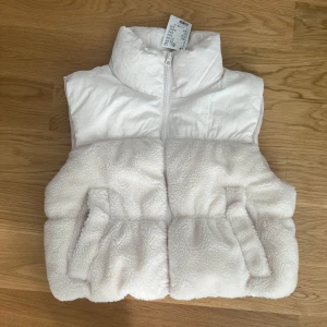 Vit och beige pälsväst från H&M - Säljer en supermysig vit och beige pälsväst från H&M. Västen har en dragkedja framtill och är perfekt för kyliga dagar. Den är helt ny aldrig använd, fick den i julklapp. Perfekt för höst och vinter!