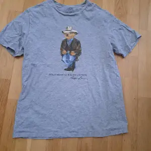 Säljer en grå t-shirt från Ralph Lauren med ett coolt tryck av en björn klädd i cowboy-outfit. T-shirten är perfekt för en avslappnad stil. Den är i okej skick med små defekter på etiketten och passar perfekt till vardags.