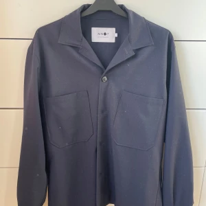 Mörkblå overshirt från NN07 - Säljer en stilren mörkblå overshirt från NN07 i storlek S. Den har en klassisk krage och två stora fickor framtill. Perfekt för lager-på-lager under höst och vår. Overshirten är i bra skick och passar både till vardags och mer uppklädda tillfällen.😃