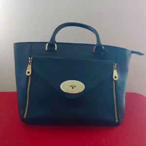 Handväska i svart skinn/kuvertväska - Väskan är sparsamt använd och i fint skick. Handtagen är 15 cm höga. Väskan har två innerfickor utan dragkedja och en större innerficka med dragkedja.Avtagbar kuvertväska. Dragkedja för stängning av väskan. Bredd: ca 43 cm,Höjd: ca 25 cm 