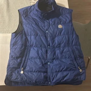 Moncler logo down vest - Storlek 5 Blå Moncler Logo down vest, självklart äkta. Cartoonen på den gick tyvvär av när jag körde moppe men annars är den i bra skick. 8/10 Som sagt självklart äkta och digital kvitto följer med.  Skriv gärna vid några funderingar!