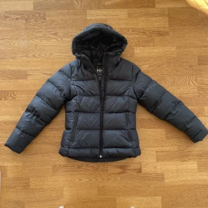 Marinblå dunjacka från Helly Hansen - Säljer en marinblå dunjacka från Helly Hansen i storlek M. Jackan är perfekt för kalla dagar med sin pufferstil och långa ärmar. Den har en praktisk dragkedja framtill och en mysig huva för extra värme. Perfekt för både höst och vinter!