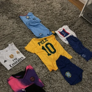 Fotbollströjor till salu - Säljer flera fotbollströjor i olika färger och stilar. Perfekt för fotbollsfans! Här finns en gul tröja med 'Pele' och nummer 10, en vit Real Madrid-tröja med Fly Emirates-logga, och en vit PSG-tröja med röd och blå detalj. Dessutom en cool rosa och svart PSG-tröja. Alla tröjor är kortärmade och i bra skick.