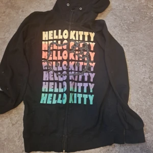 Svart Hello Kitty hoodie - Säljer en svart hoodie med Hello Kitty-tryck i regnbågsfärger. Den har en dragkedja framtill, saknar snöre till huvan. Är en barnstorlek men passar xs-s