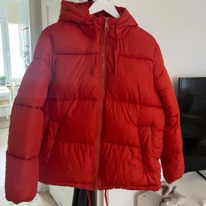 Röd/orange pufferjacka med huva - Säljer en snygg röd/orange pufferjacka i mycket bra skick. Jackan har en praktisk huva och dragkedja framtill. Perfekt för kalla vinterdagar och höstpromenader. Den är varm och bekväm.