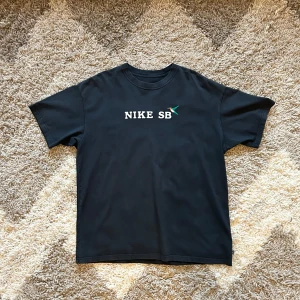 Svart t-shirt från Nike SB - Säljer en svart t-shirt från Nike SB i bomull. Den har en cool broderad fågel bredvid loggan på bröstet. Perfekt för vardagsbruk eller skate-sessioner. T-shirten är i mycket bra skick och har en normal passform. Skön och stilren design som funkar året runt!