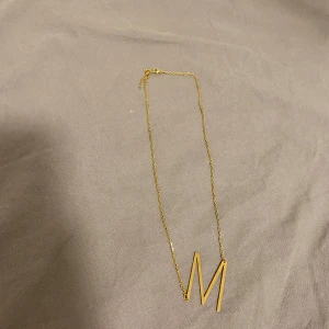 Guldhalsband med bokstaven M - Snyggt och stilrent guldhalsband med en hängande bokstav M. Perfekt för att ge en personlig touch till din outfit. Kedjan är tunn och elegant, vilket gör det lätt att bära till både vardag och fest. Ett måste för alla som älskar att bära initialer!