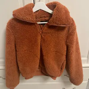 Supermysig orange teddyjacka som är perfekt för kyliga dagar. Den har en dragkedja framtill och en stor huva för extra värme. Jackan är i mycket bra skick och känns som att ha på sig ett mjukt moln. Perfekt för höst och vinter! Storlek 36