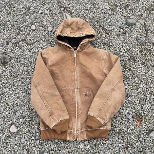 Brun jacka från Carhartt - Säljer en robust brun Carhartt jacka i mycket bra skick. Den har en praktisk huva och dragkedja framtill. Perfekt för höst och vår med sin tåliga bomullskonstruktion. Jackan har långa ärmar och muddar vid ärmslut och nederkant. Passar perfekt för casual wear! Mått är 67 cm lång och 53 cm bred, passar perfekt på mig och jag bär mest M). Buda gärna