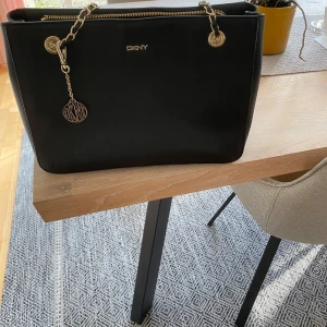 Svart axelväska från DKNY - Snygg svart axelväska från DKNY i mycket bra skick. Väskan har guldfärgade detaljer och en kedja som ger en lyxig känsla. Perfekt för både vardag och fest! 🖤