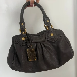 Marc Jacobs grå väska - Säljer en snygg mörkgrå handväska från Marc by Marc Jacobs i skinn. Väskan har guldfärgade detaljer och en praktisk dragkedja. Använd med super fint skick. Måtten är bredd 30cm höjd 19cm 🩶