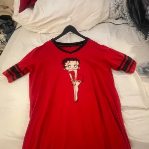 Röd Betty Boop t-shirt från Universal Studios - Säljer en cool röd t-shirt med Betty Boop-tryck från Universal Studios. Den har svarta ränder på ärmarna och nummer 30 på baksidan. Perfekt för en avslappnad stil eller som en rolig detalj i garderoben. Passar bra till jeans eller shorts!