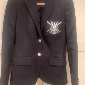 Mörkblå kavaj från Ralph Lauren - Säljer en snygg mörkblå kavaj från Ralph Lauren med broderad logga på bröstfickan. Kavajen har eleganta silverknappar och är perfekt för både formella och semi-formella tillfällen. Den är i mycket bra skick och har en klassisk passform. Perfekt för höst och vår!