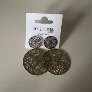 Silvriga örhängen från By Jolima - Snygga silvriga örhängen från By Jolima med ett coolt, detaljerat mönster. De har en rund form med en glittrig yta på den övre delen och ett större, utskuret mönster på den nedre delen. Perfekta för att ge lite extra bling till din outfit! Helt nya!!