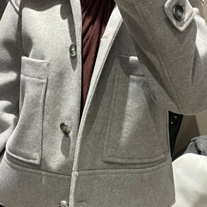 Grå kavaj med knappar - Snygg grå kavaj i ull med stora knappar fram och på ärmarna. för höst och vår, med en stilren look som passar både till vardags och fest. Två fickor fram för extra stil och funktionalitet.
