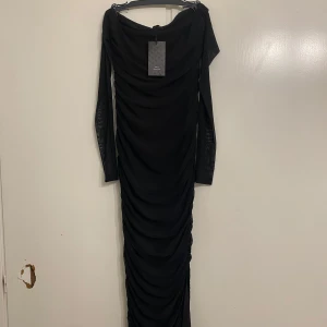 Svart långklänning med meshärmar - Elegant svart långklänning med långa meshärmar och rynkad design. Perfekt för festliga tillfällen. Klänningen har en tight passform som framhäver figuren.