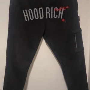 Svarta mjukisbyxor från Hoodrich - Säljer ett par svarta mjukisbyxor från Hoodrich i storlek S. Byxorna har en cool design med 'Hoodrich' broderat i vitt och rött på framsidan. Perfekta för en avslappnad stil och bekväma att bära. Passar bra till vardags eller chillkvällar.finns en liten fläck fram vid texten inget som direkt stör men den finns där 