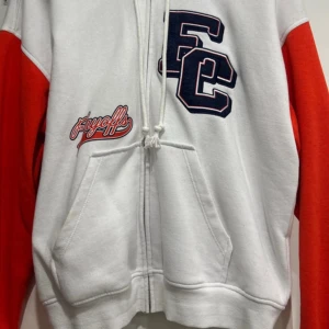 Vit och orange hoodie med tryck - Säljer en cool vit hoodie med orange ärmar och tryck på bröstet och ärmen. Perfekt för en avslappnad stil. Den har en dragkedja framtill och en stor ficka. Passar perfekt för höst och vår!