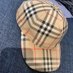 Rutig keps från Burberry - Säljer en snygg rutig keps från Burberry i klassiskt beige, svart och rött mönster. Kepsen har justerbar passform och är perfekt för både vardag och fest. Märkeslogga på baksidan ger en extra touch av stil. Passar perfekt till alla säsonger!