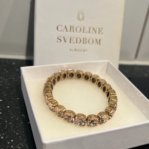 Guldarmband från Caroline Svedbom - Snyggt guldarmband från Caroline Svedbom med stora, glittrande stenar. Perfekt för att ge en touch av glamour till vilken outfit som helst. Armbandet är elastiskt och passar de flesta handleder. Kommer i originalförpackning.