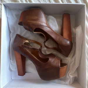 Bruna klackskor från Jeffrey Campbell - Säljer ett par snygga bruna klackskor från Jeffrey Campbell i modellen Foxy Wood, storlek 39. De har en hög, stabil klack och en peep-toe design. Skorna är i skinn och har en rem över foten för extra stöd. Knappt använda och kommer med originalbox samt damm/skyddspåsar. Perfekta för en utekväll eller fest! 🥿✨