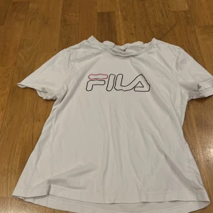 Vit t-shirt från FILA - Säljer en vit t-shirt från FILA i storlek L, men mer som en M. Den har en klassisk design med korta ärmar och FILA-loggan tryckt på framsidan i svart och rött. Perfekt för en avslappnad stil eller träning. T-shirten är i bra skick, dock har ena sömnen på armen gått sönder.