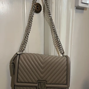 Victoriasecret väska - Snygg beige axelväska med quiltat mönster och silverkedja. Perfekt för både vardag och fest! Väskan har en stilren design med ett V-emblem på framsidan.