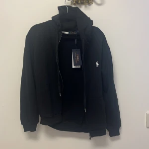 Svart hoodie från Ralph Lauren - Säljer en stilren svart hoodie från Ralph Lauren i mycket bra skick. Den har en dragkedja framtill och det klassiska logotypen broderad på bröstet. Perfekt för både vardag och chillkvällar. Passar till allt och är superbekväm!