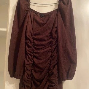 Brun klänning med rynkdetaljer - Säljer en snygg brun klänning med rynkdetaljer framtill. Den har långa ärmar och en loose passform, perfekt för höst och vinter. Materialet känns mjukt och bekvämt, vilket gör den idealisk för både vardag och festligare tillfällen.