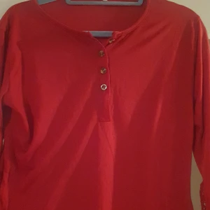 Röd långärmad topp med spetsdetaljer - Säljer en snygg röd långärmad topp med knappar framtill och vackra spetsdetaljer längs ärmarna. Perfekt för både vardag och festliga tillfällen.  Passar perfekt till höst- och vårgarderoben!