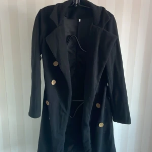 Svart kappa /jacka med knappar - Säljer en stilren svart kappa/jacka  i storlek S. Den har långa ärmar och snygga guldknappar framtill. Perfekt för höst och vinter, och funkar både till vardags och fest. Kappan är i bra skick och ger en elegant look.