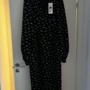 Svart blommig klänning från ADOORE - Snygg svart klänning från ADOORE med ett gulligt blommönster. Den har långa ärmar och en dragkedja i ryggen. Perfekt för både vardag och festliga tillfällen. Klänningen är i nytt skick med taggen kvar.