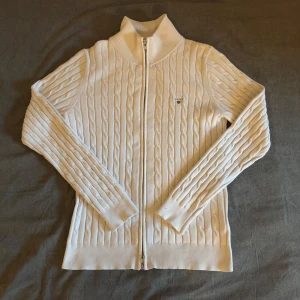 Vit kabelstickad kofta med dragkedja - Säljer en stilren vit kofta med dragkedja från gant. Koftan är i mycket bra skick förutom en liten fläck på vänstra armen. 