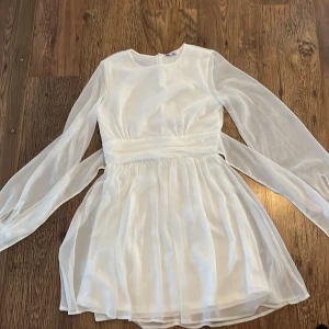 Vit klänning med knytband - Säljer en superfin vit klänning med långa ärmar och ett elegant knytband i midjan. Klänningen har en luftig och flödande design, perfekt för sommarens alla fester. Materialet känns lätt och bekvämt mot huden. Passar perfekt för en stilren och chic look!