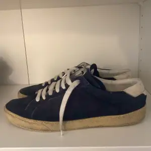 Säljer ett par snygga blå sneakers från Saint Laurent i mocka. De har vita snören och en vit häl med logotyp. Sulan är i en beige ton och ger en cool vintage-look. Perfekta för både vardag och fest!