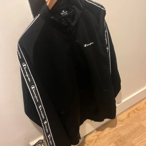 Svart jacka från Champion - Säljer en svart Champion jacka i storlek XL. Den har långa ärmar med Champion-loggan längs sidorna och en dragkedja framtill. Perfekt för höst och vår, och den är i mycket bra skick. Passar både till träning och vardagsbruk. Pris kan diskuteras