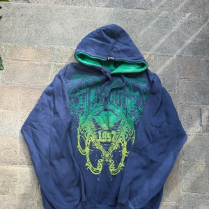 Blå Tapout Hoodie - Säljer en cool blå/lila hoodie från Tapout med grönt/gult tryck och grön insida av huvan. Perfekt för träning eller chill. Den har en stor logga på framsidan och en mindre på baksidan. Hoodien är i bra skick och superbekväm. Passar perfekt för höst och vår! Tröjan är i nyskick men har använts.