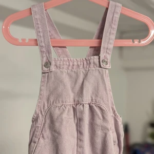 Ljusrosa hängselbyxor från Zara, strl 98 - Super gulliga ljusrosa hängselbyxor från Zara för barn i storlek 2-3 år. De är gjorda i ett mjukt bomullsmaterial och har justerbara axelband med knappar.