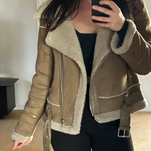 ZARA- Beige mockajacka med foder - Säljer en superfin beige mockajacka med mysigt foder. Jackan har en cool bikerstil med dragkedjor och ett bälte i midjan. Perfekt för höst och vår när du vill hålla dig varm och snygg. Den har långa ärmar och en skön passform. Jackan är använd men I mycket bra skick tack vare hög kvalitet och slitstarka material.